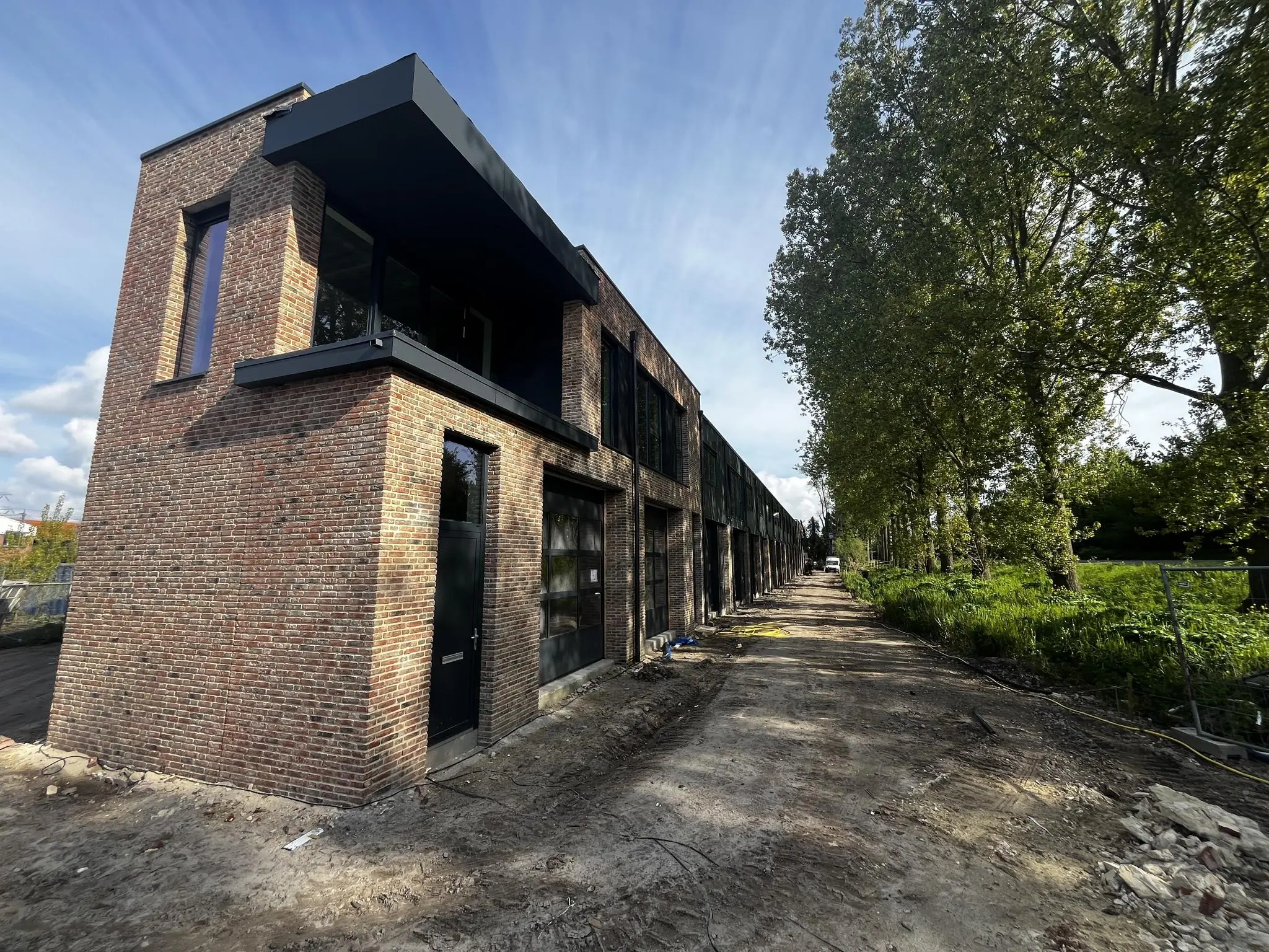 Nieuwbouw van moderne rijwoningen aan de Broekdijk West met een onverharde weg en hoge bomen ernaast.